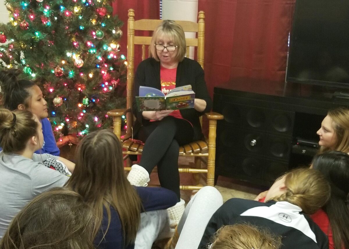 Houseparent Sandy Pape sits in a rocking chair, holding a book. She is beside a Christmas tree. Girls sit at her feet in a circle.